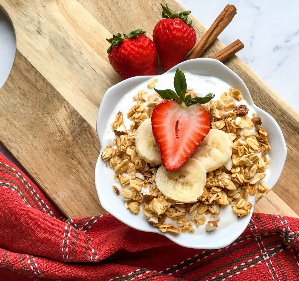 A bowl full of homemade Greek Yogurt topped with granola, bananas, and apples on a wooden board