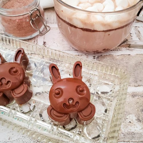 Two bunny faces made out of chocolate next to a glass mug of hot chocolate on a glass tray
