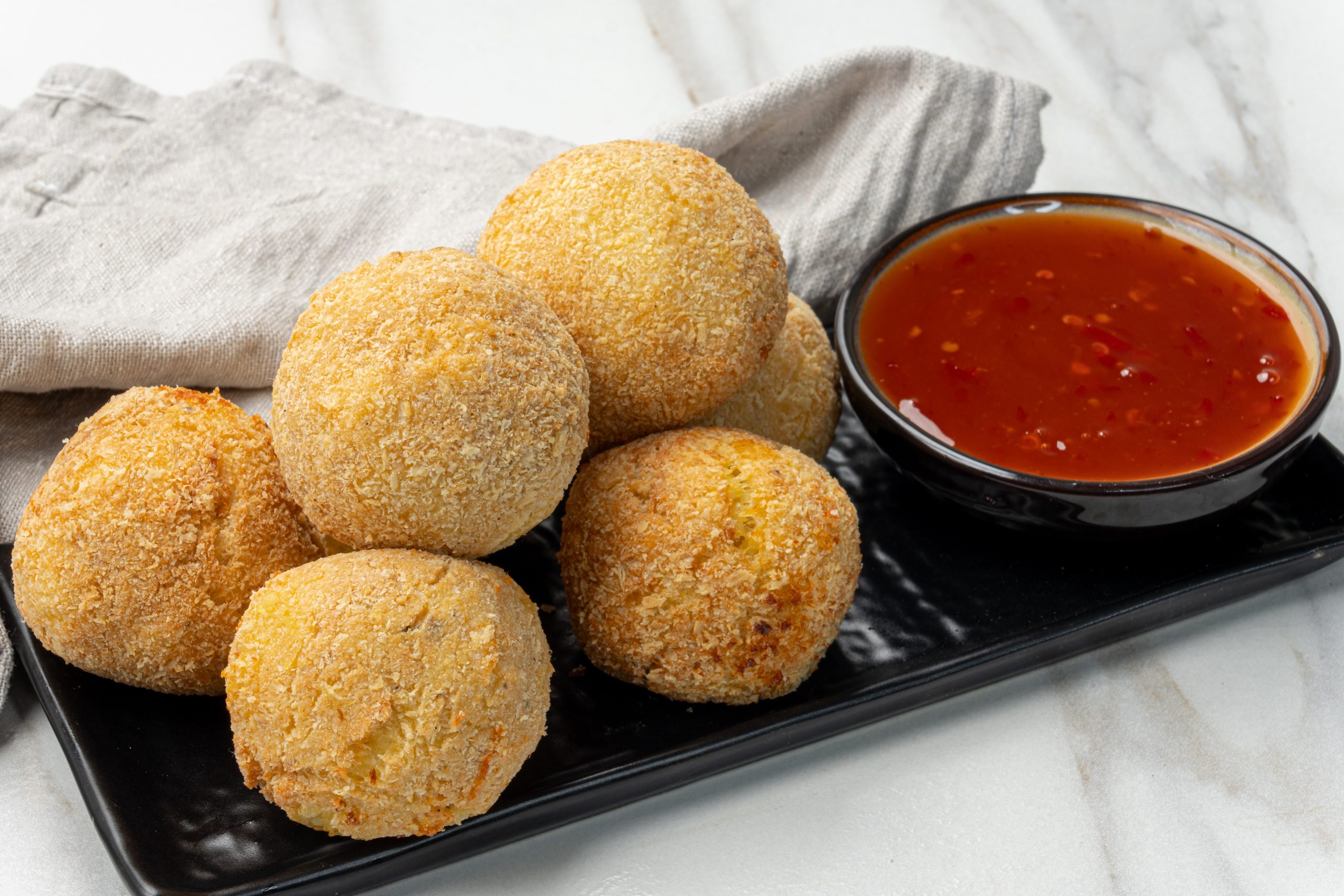 Golden brown potato cheese croquettes coated in breadcrumbs, served on a black plate with a bowl of dipping sauce.