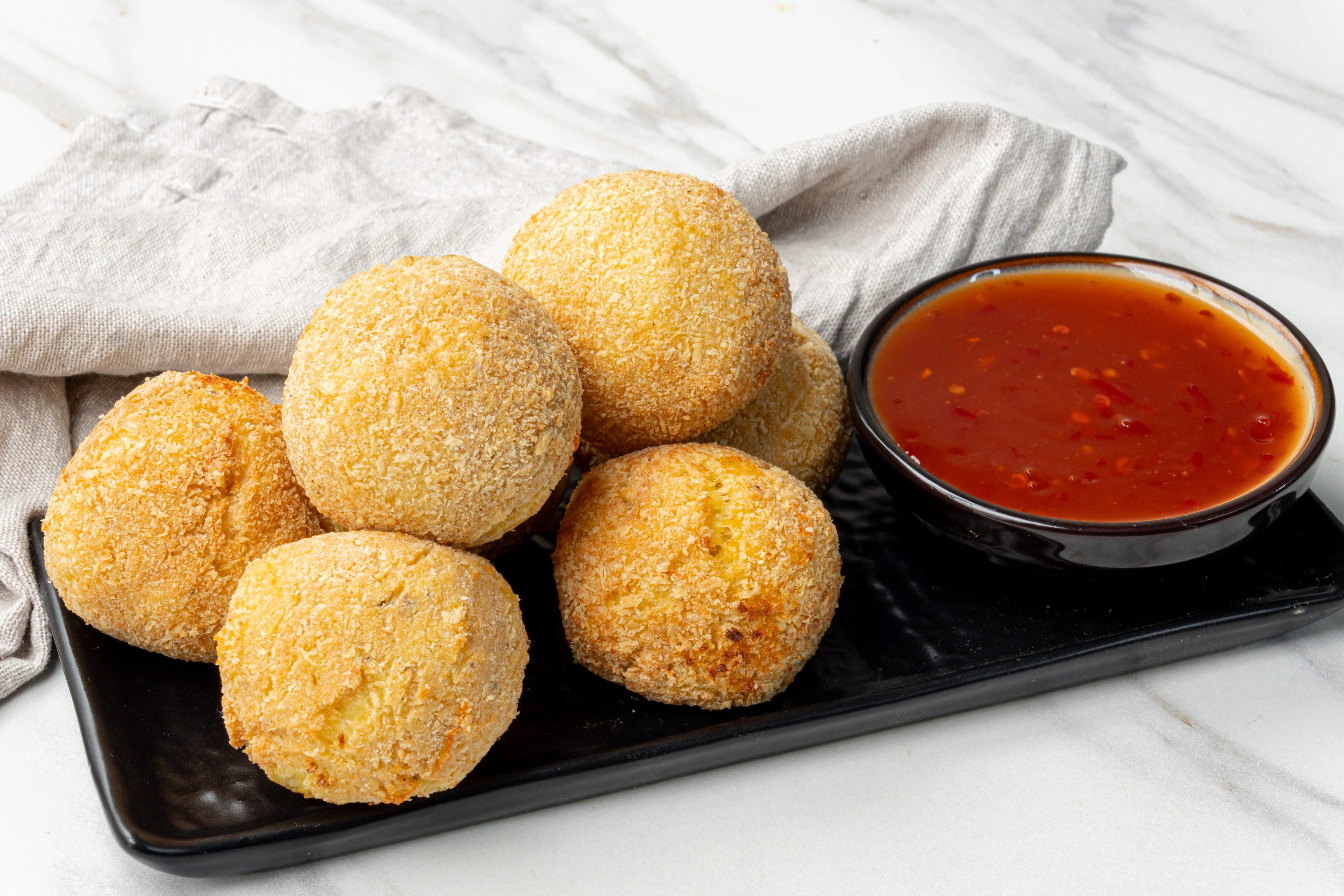 Golden brown potato cheese croquettes coated in breadcrumbs, served on a black plate with a bowl of dipping sauce.