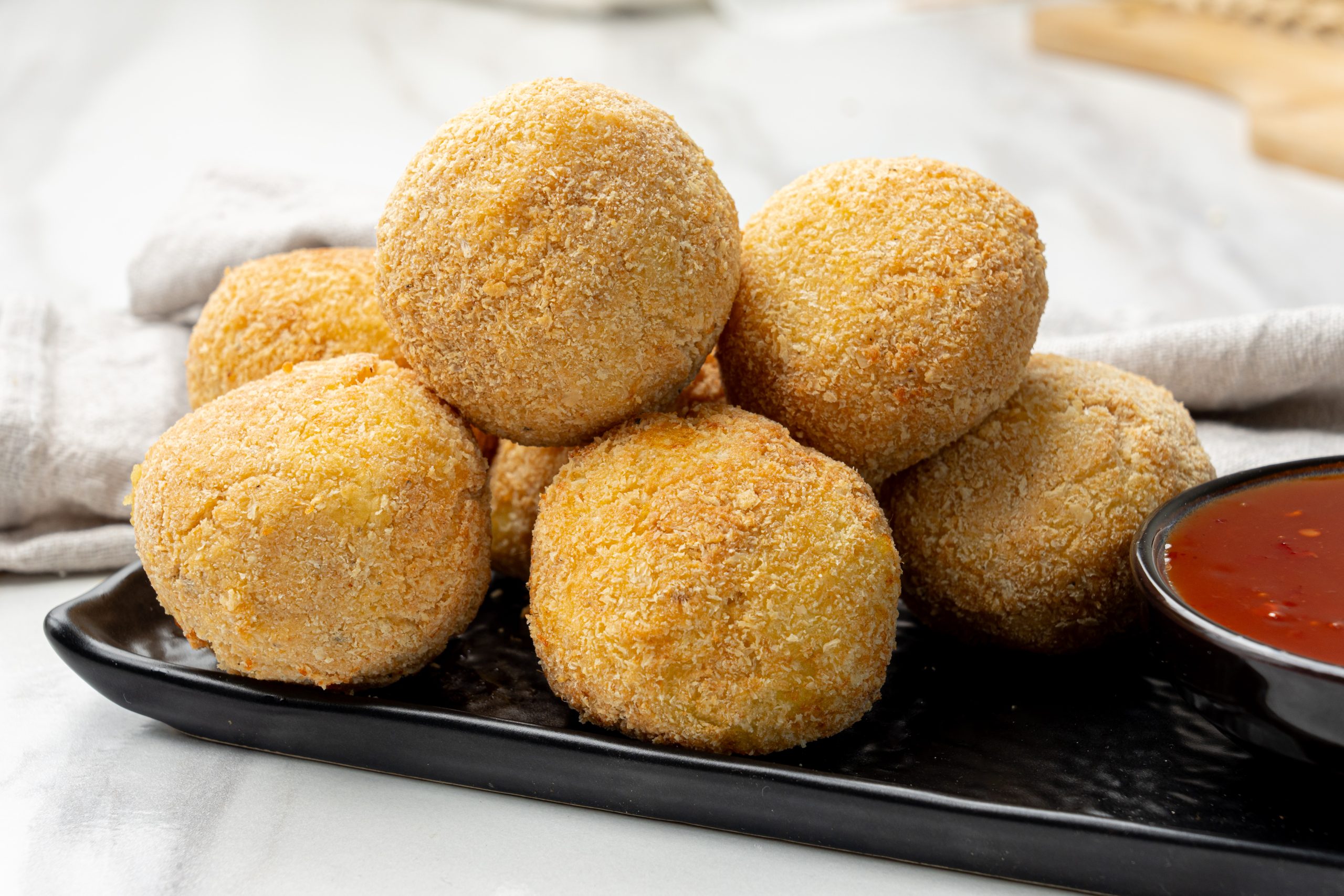 Golden brown potato cheese croquettes coated in breadcrumbs, served on a black plate with a bowl of dipping sauce.