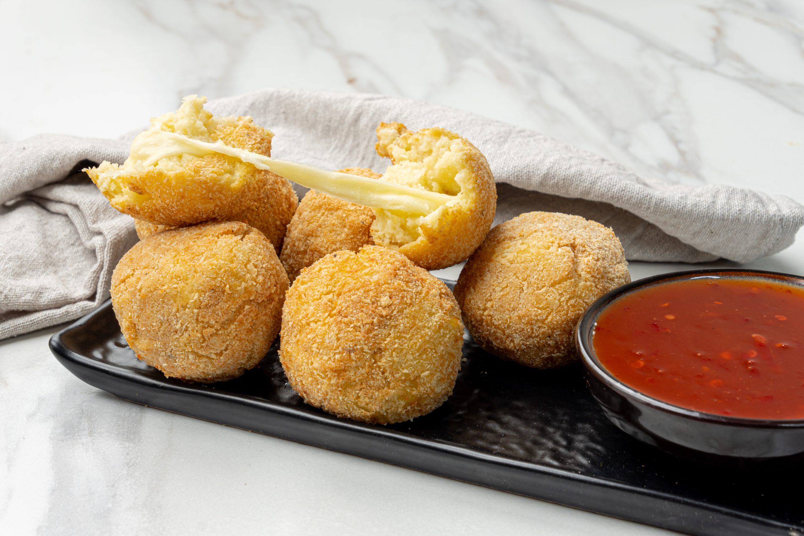 Golden brown potato cheese croquettes coated in breadcrumbs, served on a black plate with a bowl of dipping sauce.