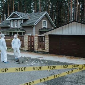 Crime scene investigators in protective suits examining evidence at a suburban house with yellow tape.