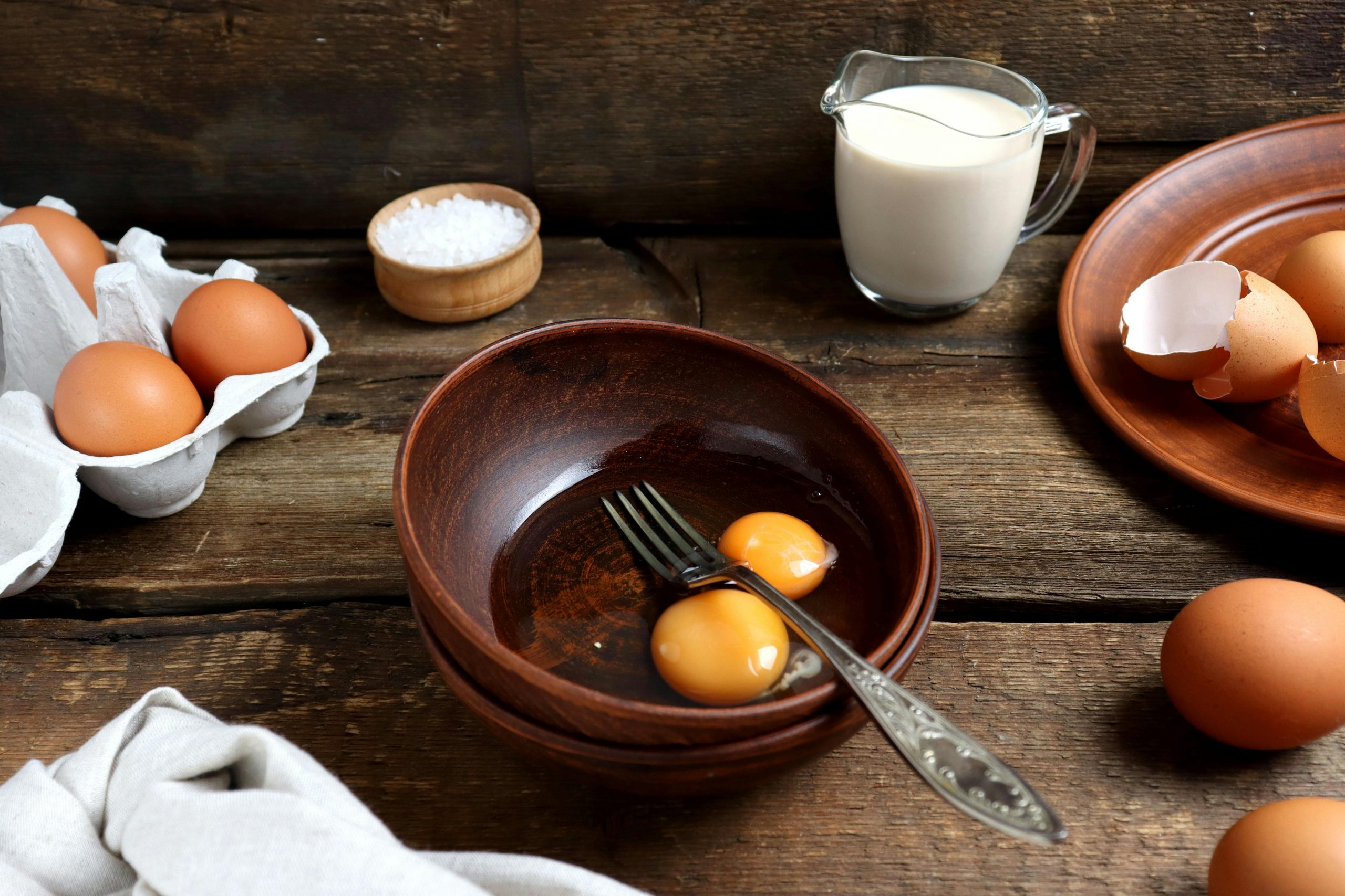 Still life photograph featuring eggs, milk, and kitchenware on a rustic wooden table.