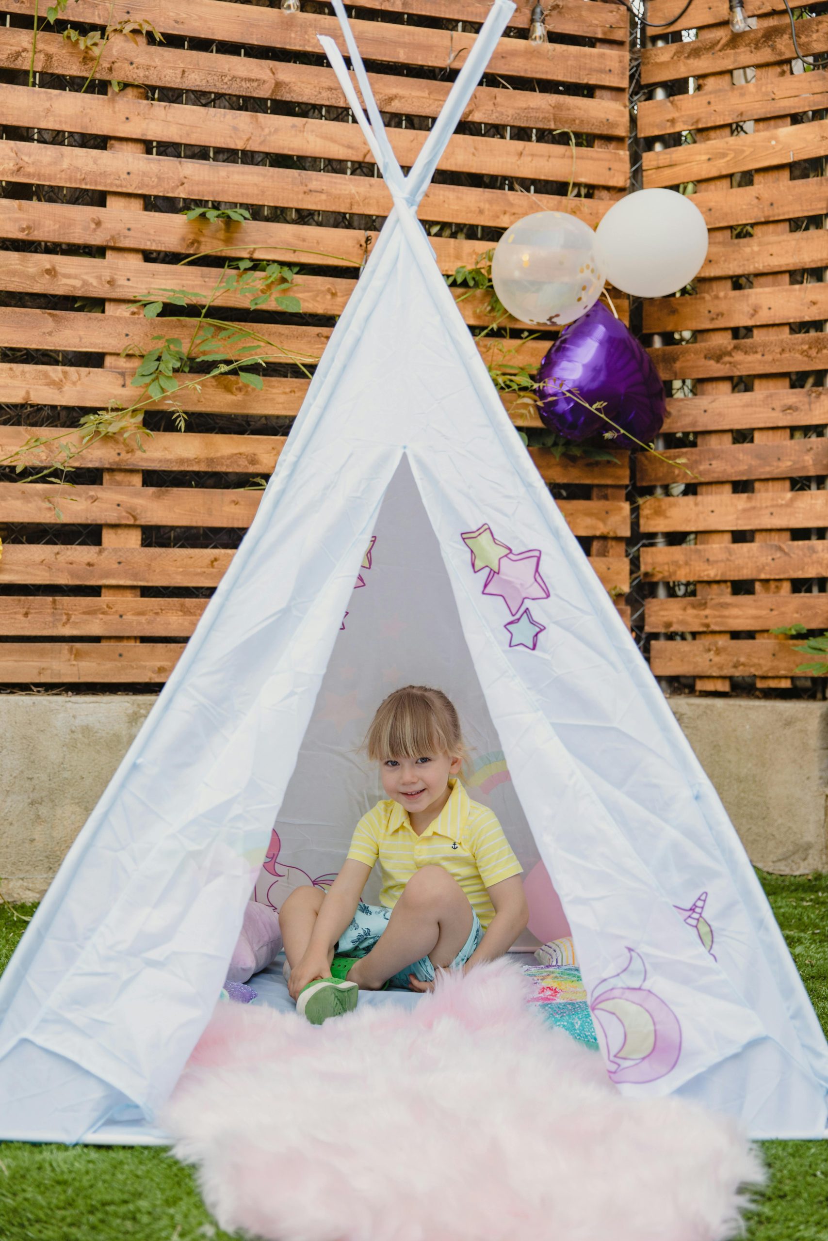 Adorable child in a teepee tent with colorful balloons in a backyard setting.
