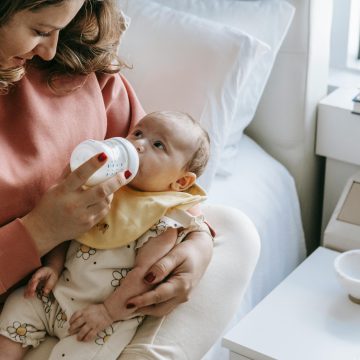 From above of crop happy young mother in casual clothes smiling while hugging and feeding adorable newborn from bottle sitting on comfortable bed