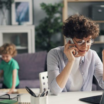 A woman multitasking with a phone and laptop while her child plays in the background.