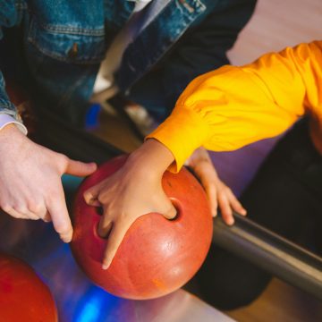 Close-up of hands preparing a red bowling ball in a vibrant bowling alley setting.