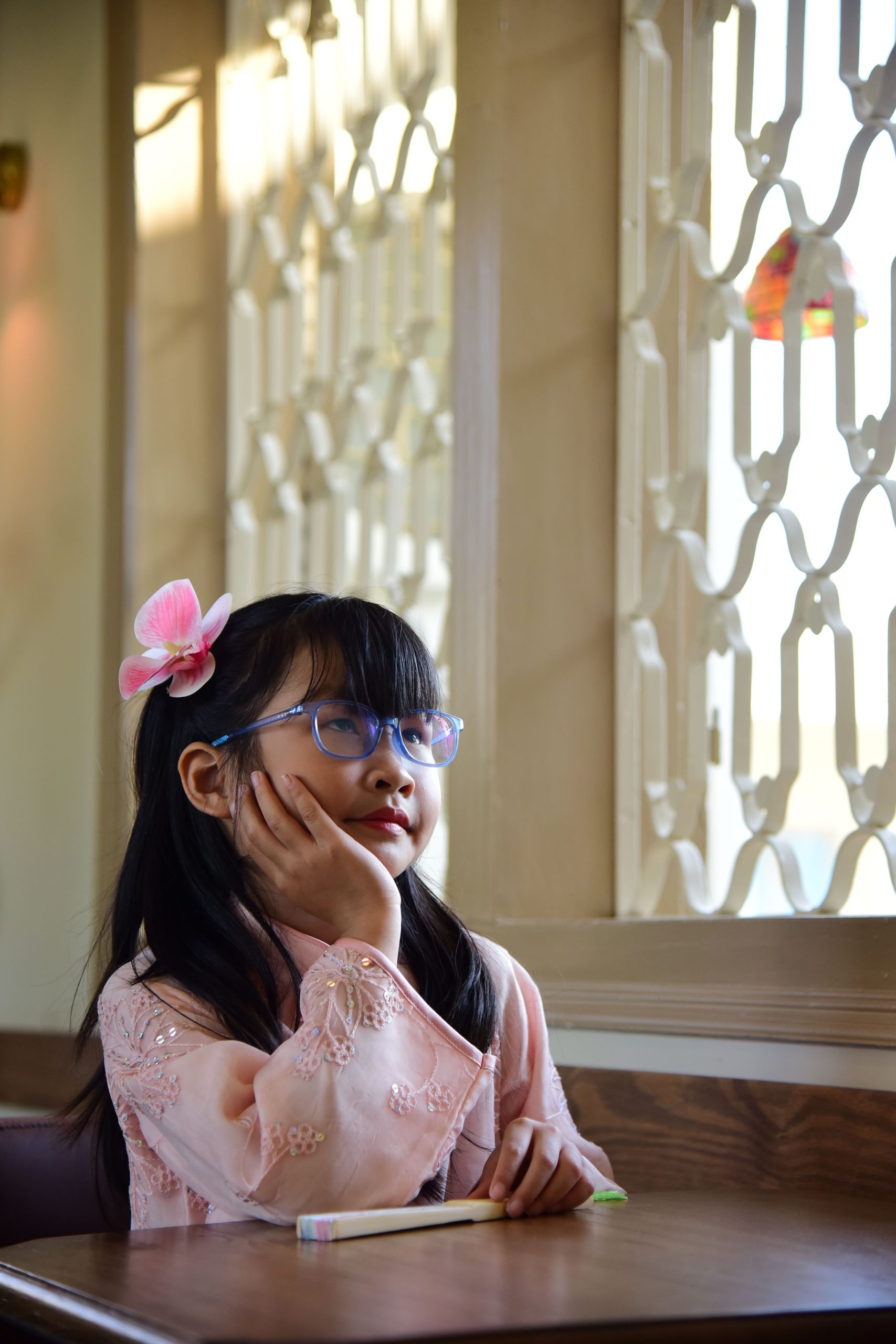 A young girl with glasses and a flower hair clip looks thoughtful indoors in natural light.