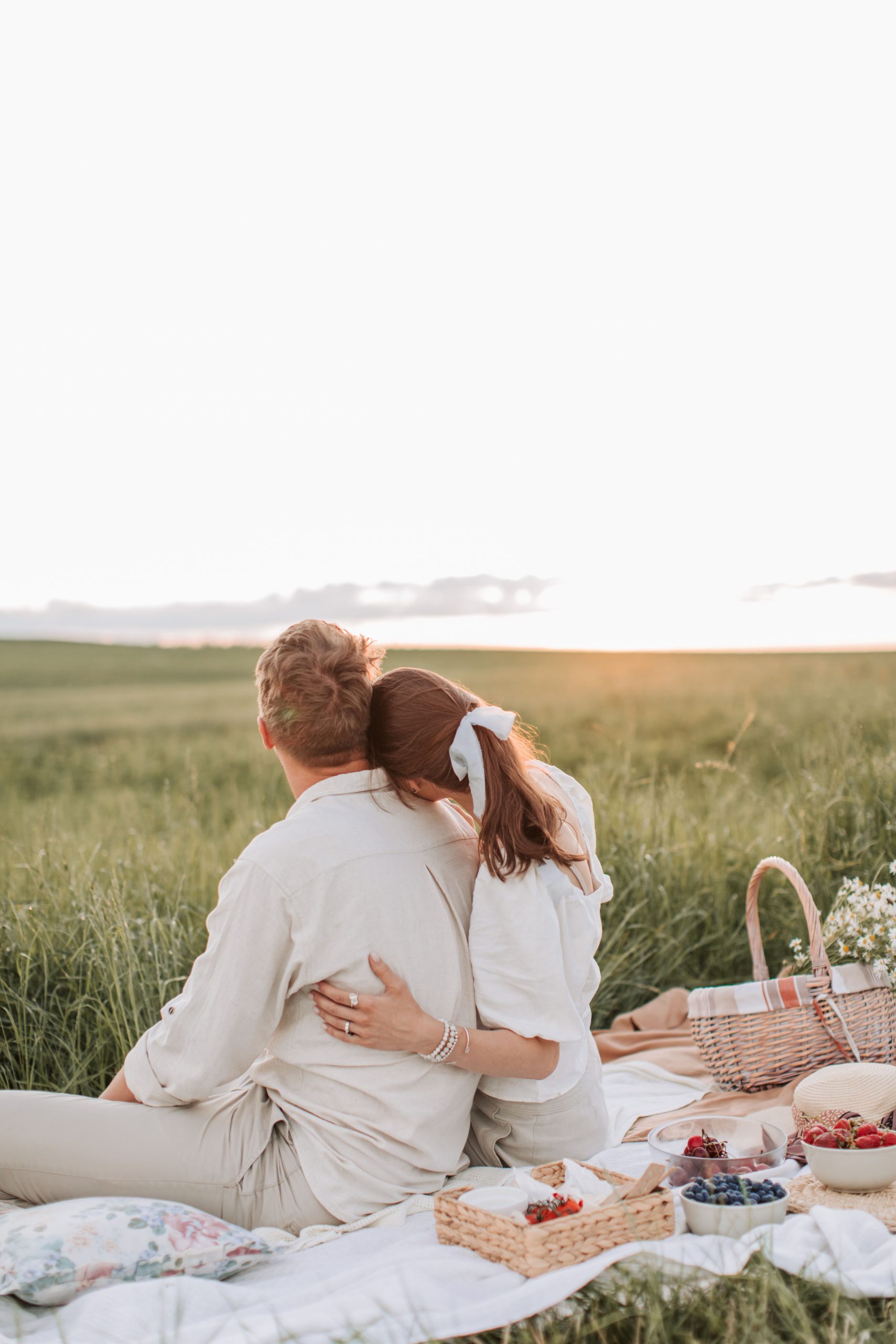 Couple enjoying a romantic sunset picnic in a grassy field with a variety of fruits and a cozy blanket.