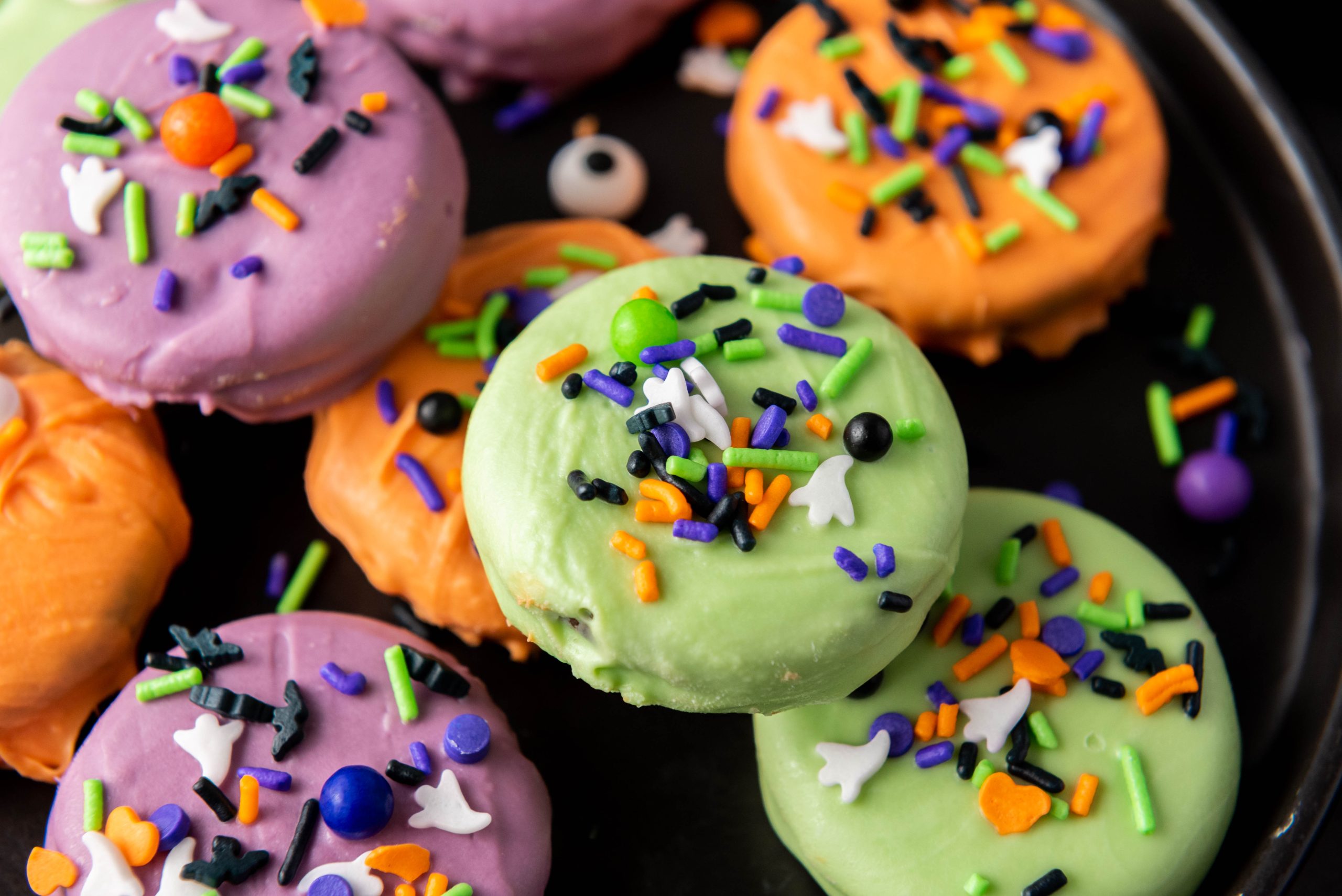 Colorful Halloween Oreos dipped in orange, purple, and green candy melts and topped with festive sprinkles and candy eyes.