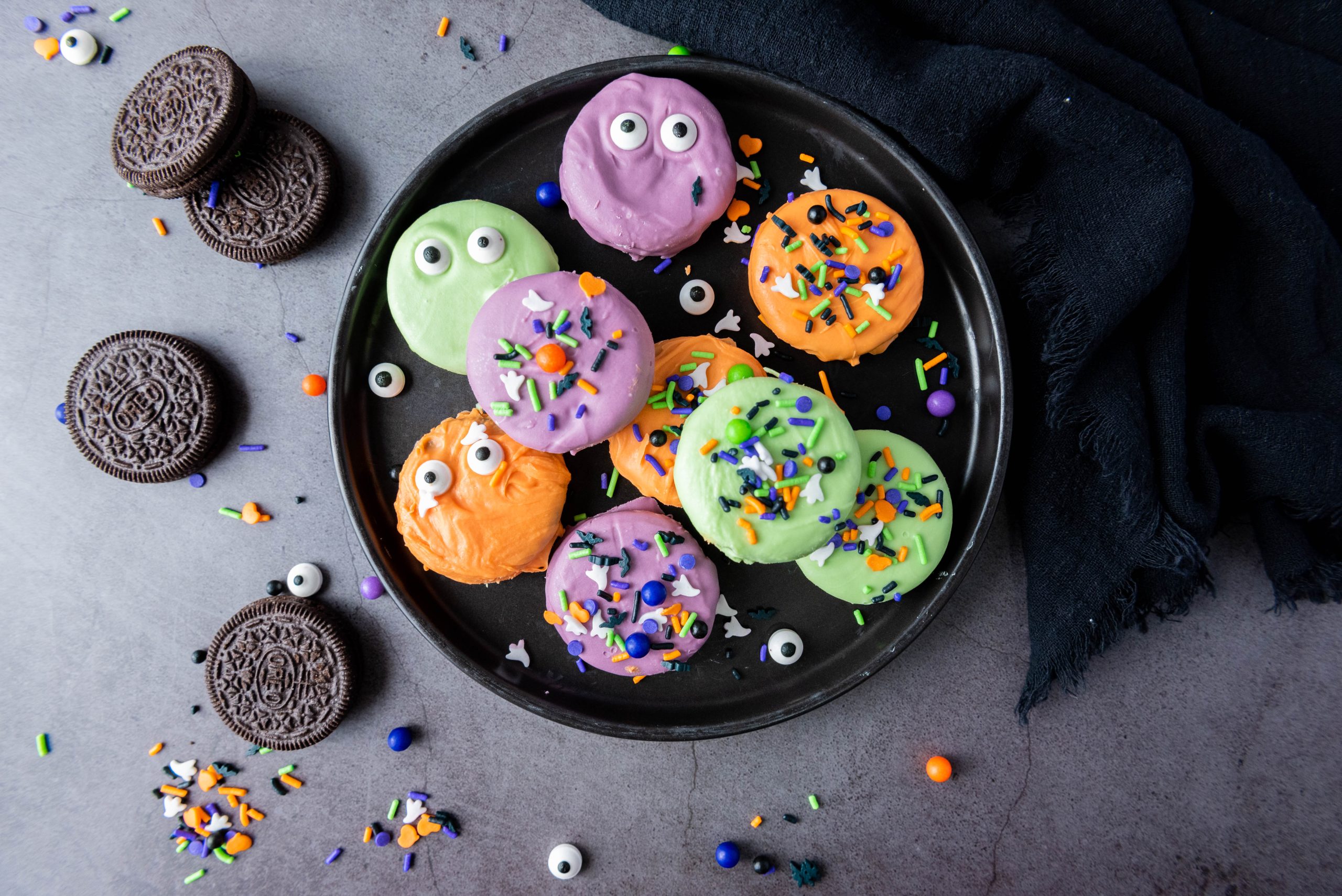 Colorful Halloween Oreos coated in orange, purple, and green candy melts with spooky sprinkles and candy eyes on a black serving tray.