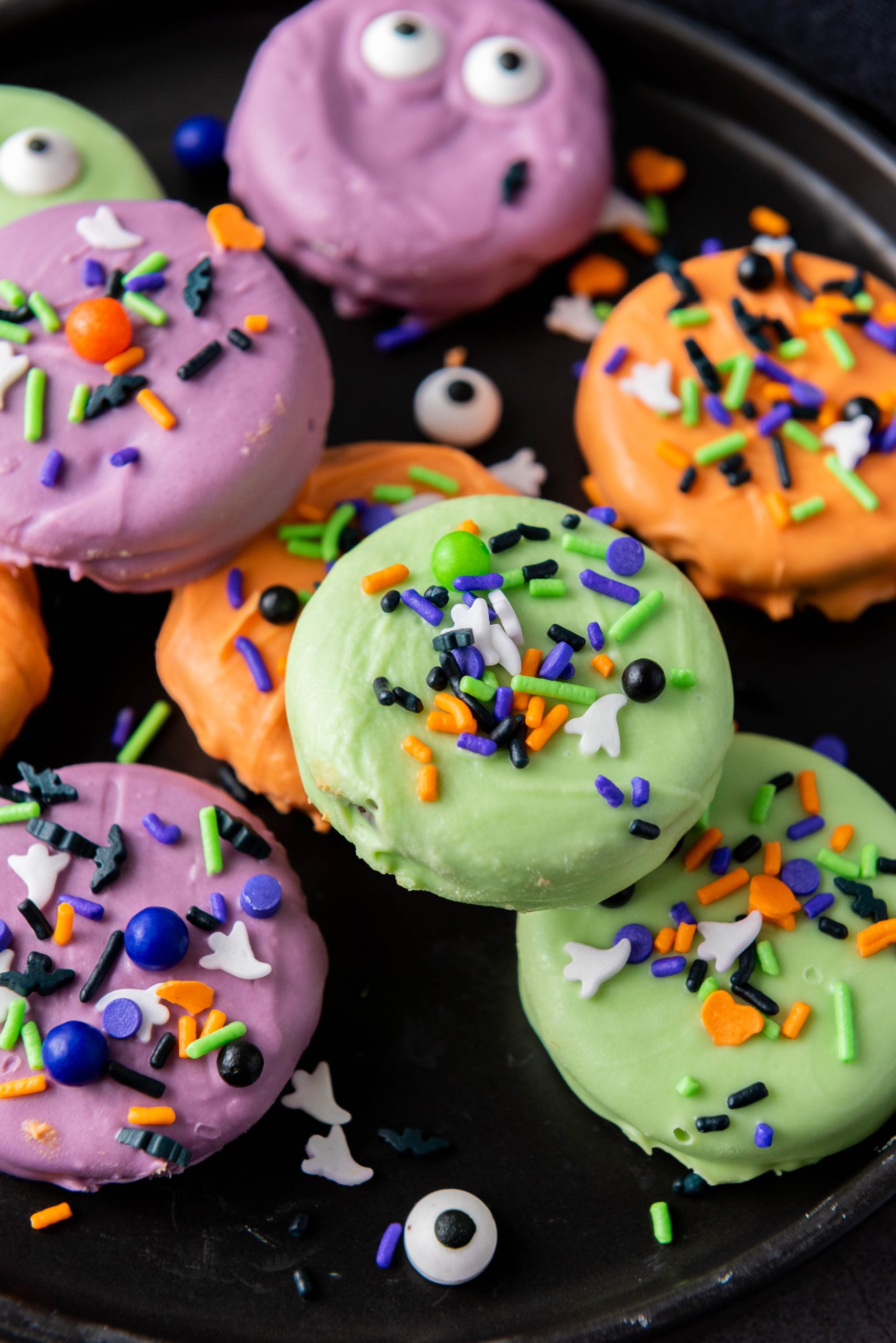 Colorful Halloween Oreos decorated with orange, purple, and green candy melts, candy eyes, and festive Halloween sprinkles.