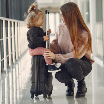 Full length of positive content woman and cute stylish blond little girl sitting on black suitcase in airport corridor while waiting for flight