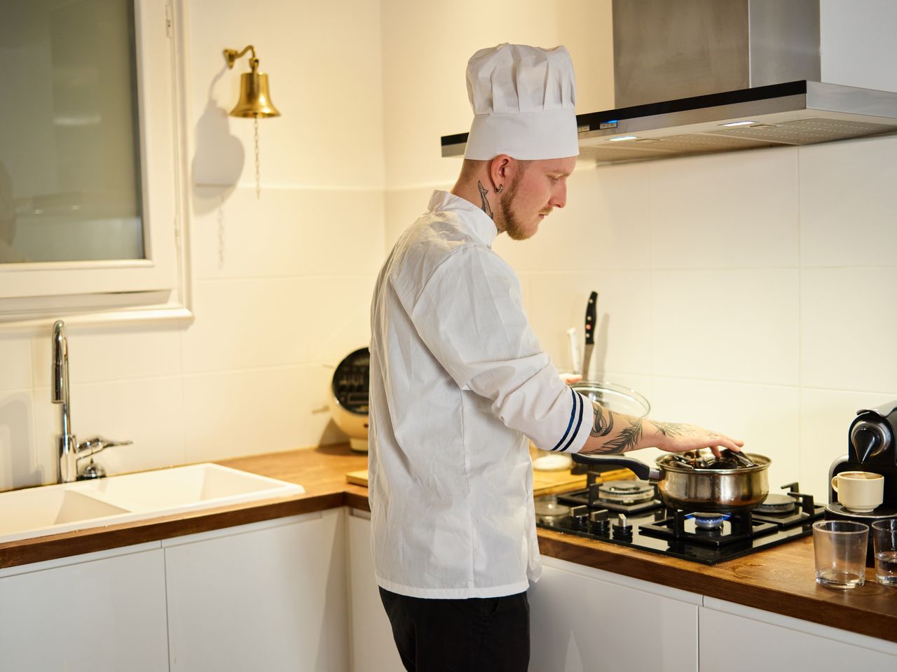 A tattooed chef wearing a hat cooks in a modern kitchen, focusing on a pot on the stove.